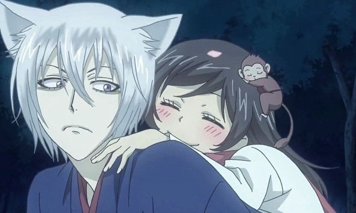 Tomoe with white fox ears and silver hair carrying Nanami Momozono on his back while she smiles with a small monkey sitting on her head, in a night scene from the anime Kamisama Kiss.